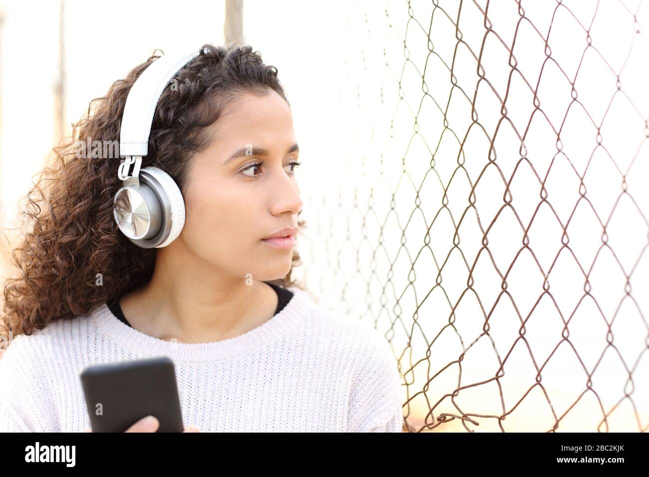 La ragazza seria della latina che ascolta la musica e tiene il telefono intelligente nella strada guarda via attraverso una griglia Foto Stock