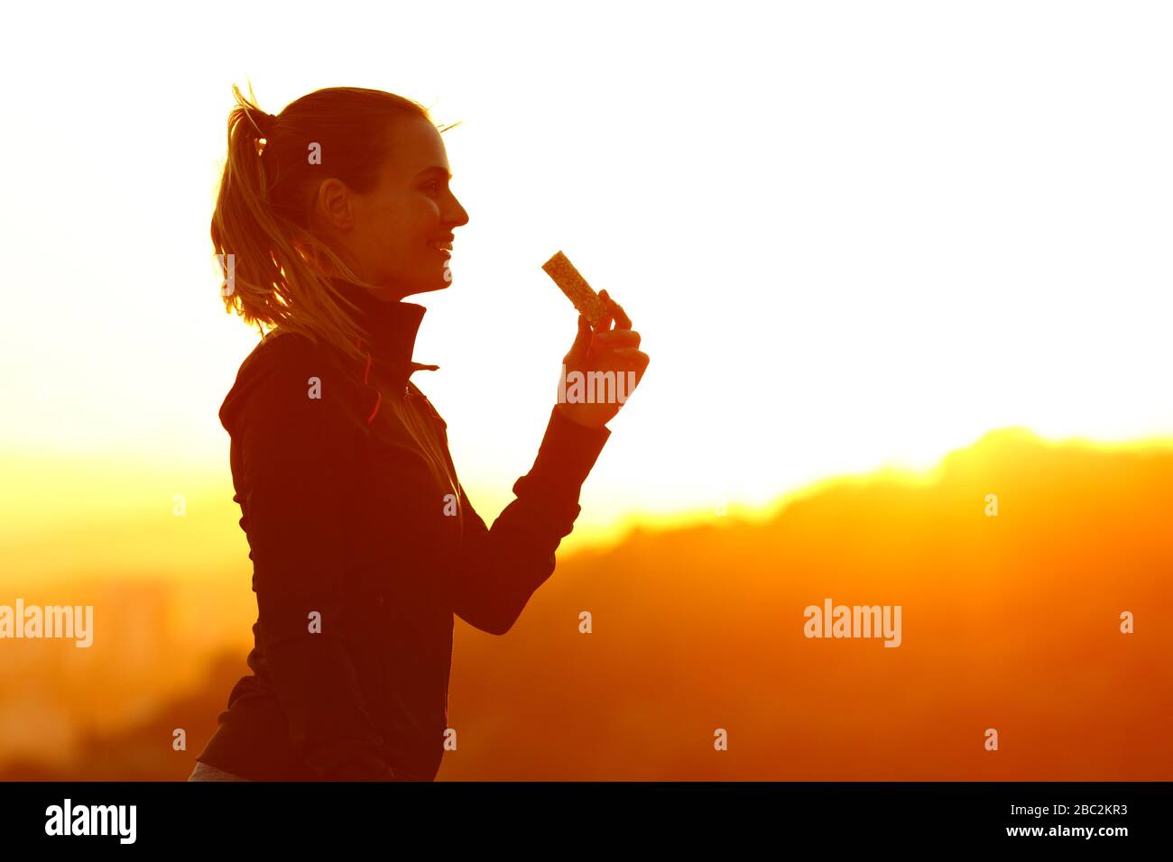 Silhouette di runner donna mangiare energia bar dopo aver correre al tramonto in montagna Foto Stock