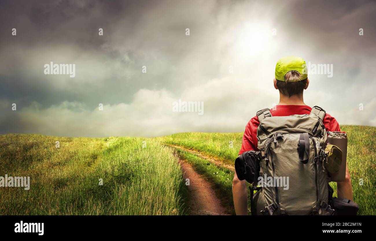 Giovane uomo che fa trekking un sentiero che conduce all'orizzonte sulla campagna aperta, con cielo drammatico e l'umore chiaro Foto Stock