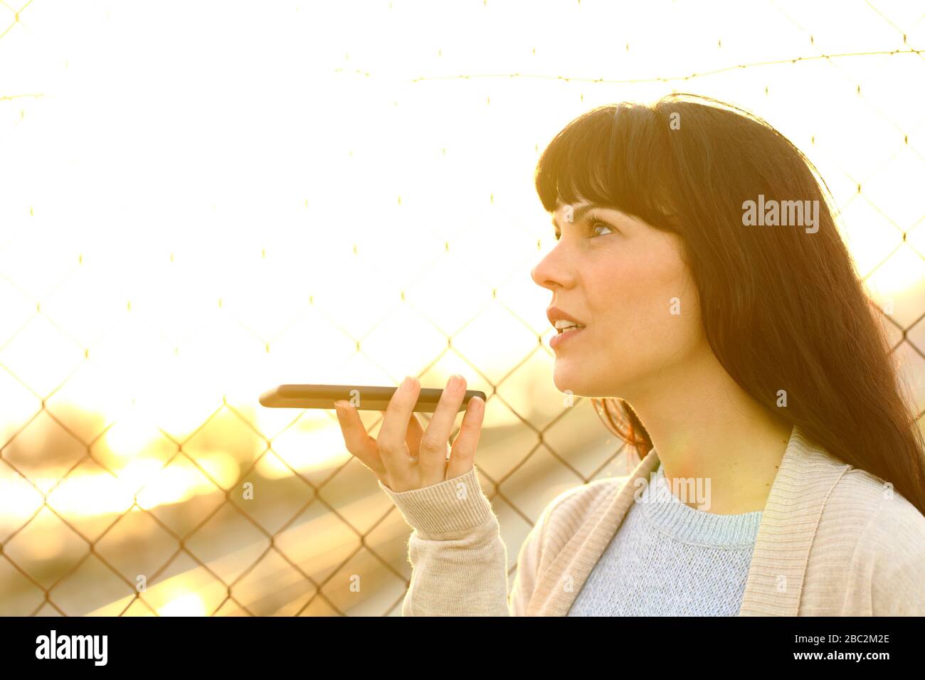 Donna adulta che parla registrare il messaggio utilizzando il riconoscimento vocale sul telefono cellulare in strada al tramonto Foto Stock