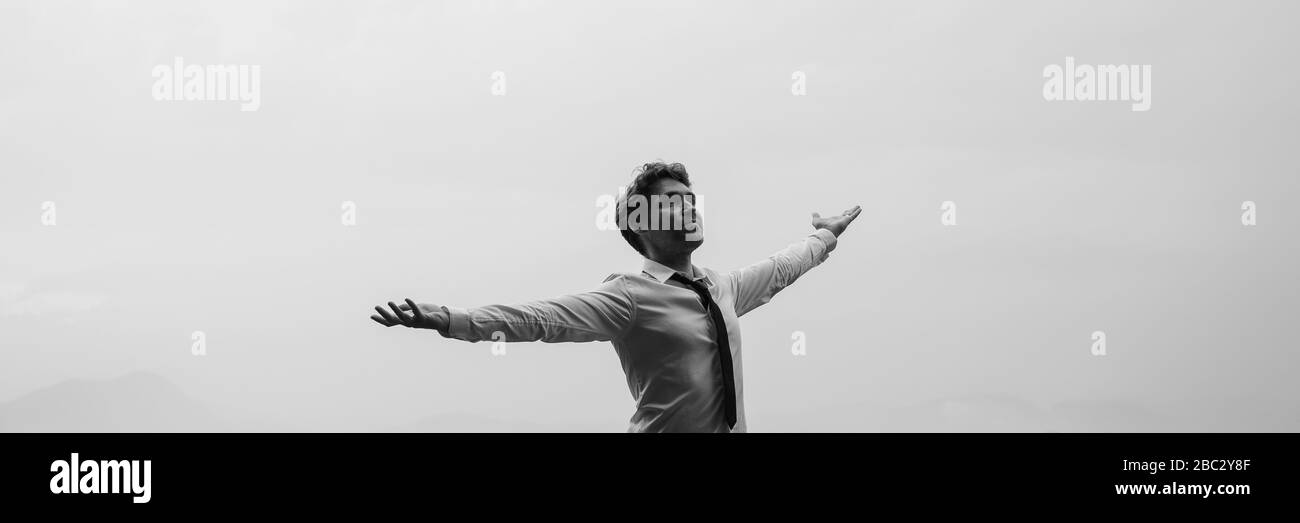 Immagine a scala di grigi di un uomo d'affari che si trova sotto il cielo nuvoloso con le braccia spalancate. Foto Stock