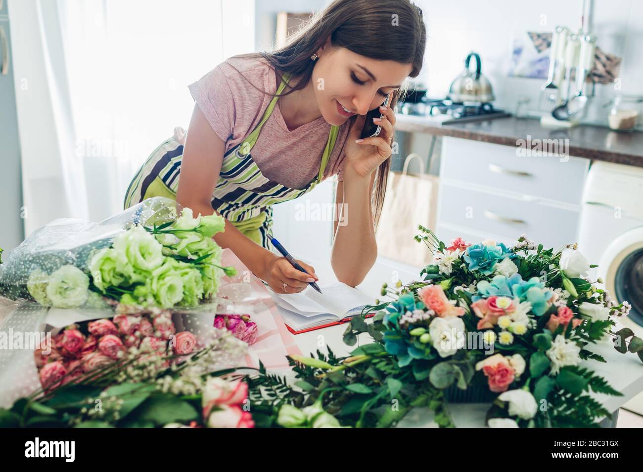 Lavoro da casa durante la quarantena del coronavirus. Donna fiorista compone bouquet di fiori riceve ordini al telefono. Consegna Foto Stock