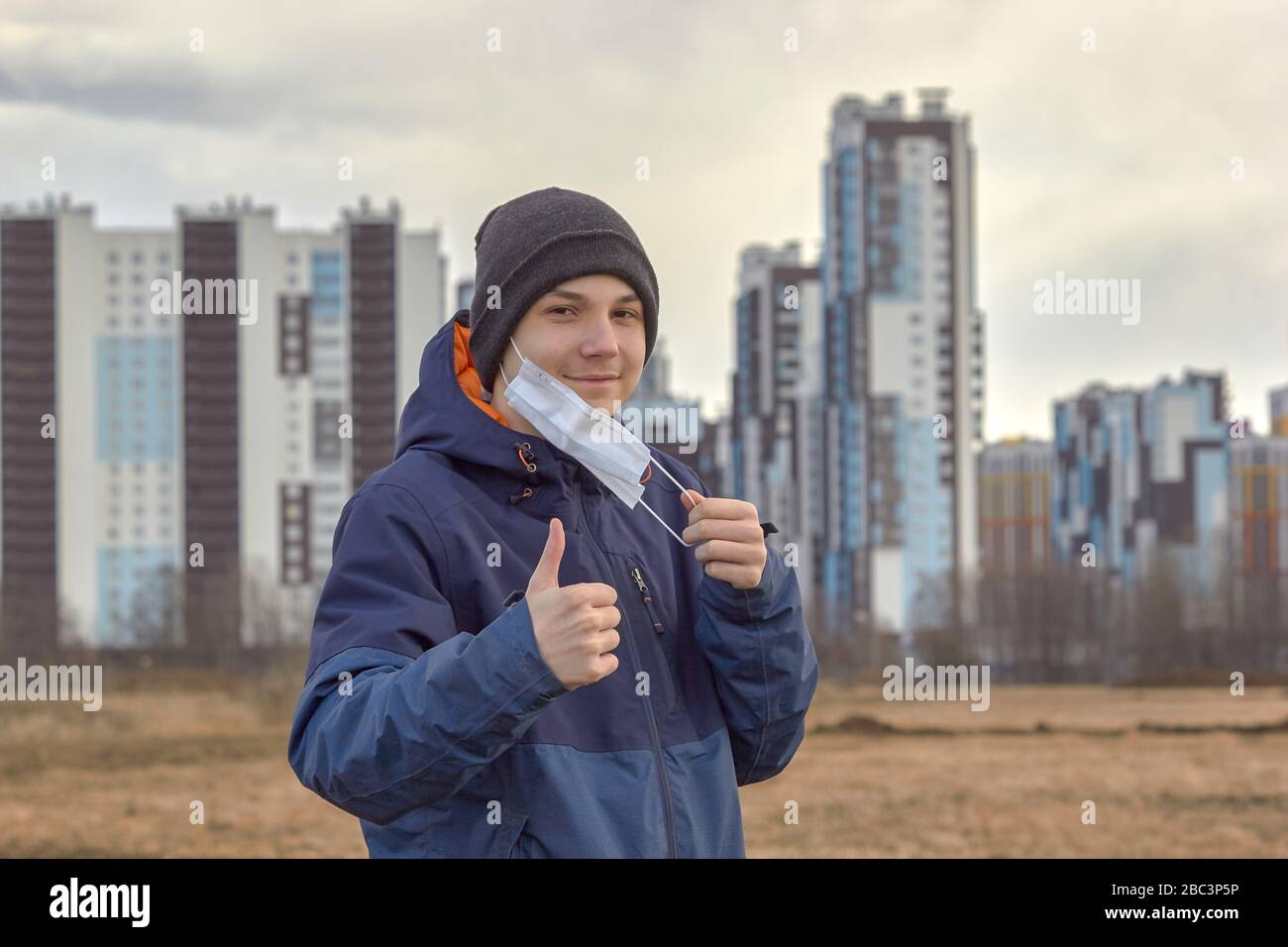 un adolescente in abiti scuri demi-stagione toglie una maschera medica contro lo sfondo di alti blocchi Foto Stock
