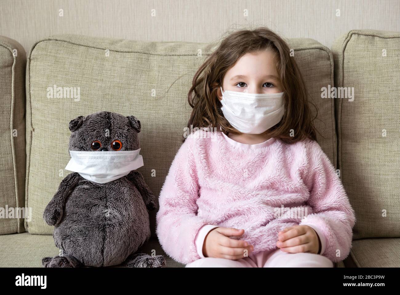 Concetto di quarantena per coronavirus, bambina con giocattolo sul divano a casa durante la pandemia di COVID-19. Capretto in maschera medica per la protezione al virus corona ind Foto Stock
