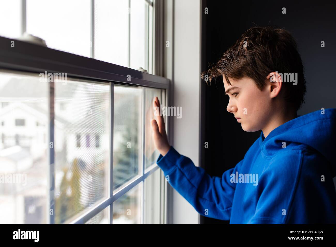 Tween ragazzo che guarda fuori da una finestra in una stanza buia. Foto Stock