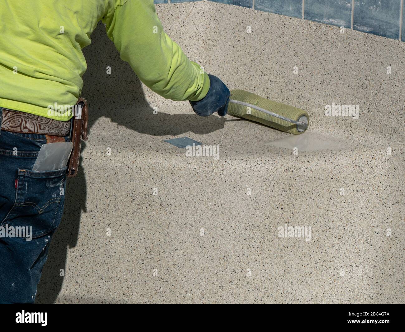 Uomo che usa un rullo per rimuovere l'acqua in eccesso dal cemento appena applicato della piscina contenente ciottoli Foto Stock