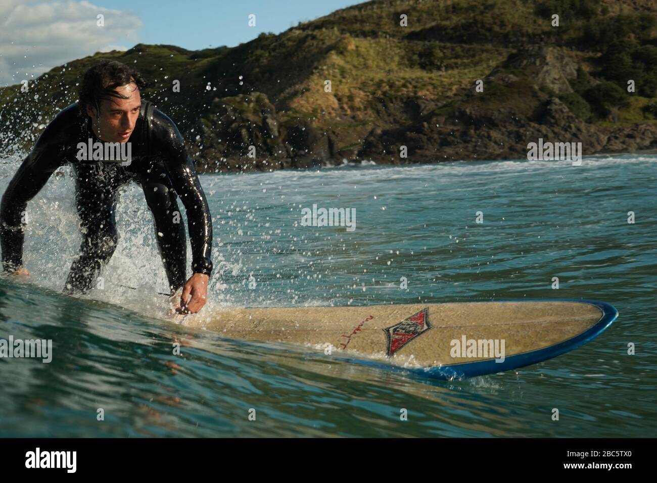 Un giovane surfista maschile in una muta nera guida un longboard surf su un'onda di rottura con acqua bianca. Giorno d'estate con un cielo blu nuvoloso. Foto Stock