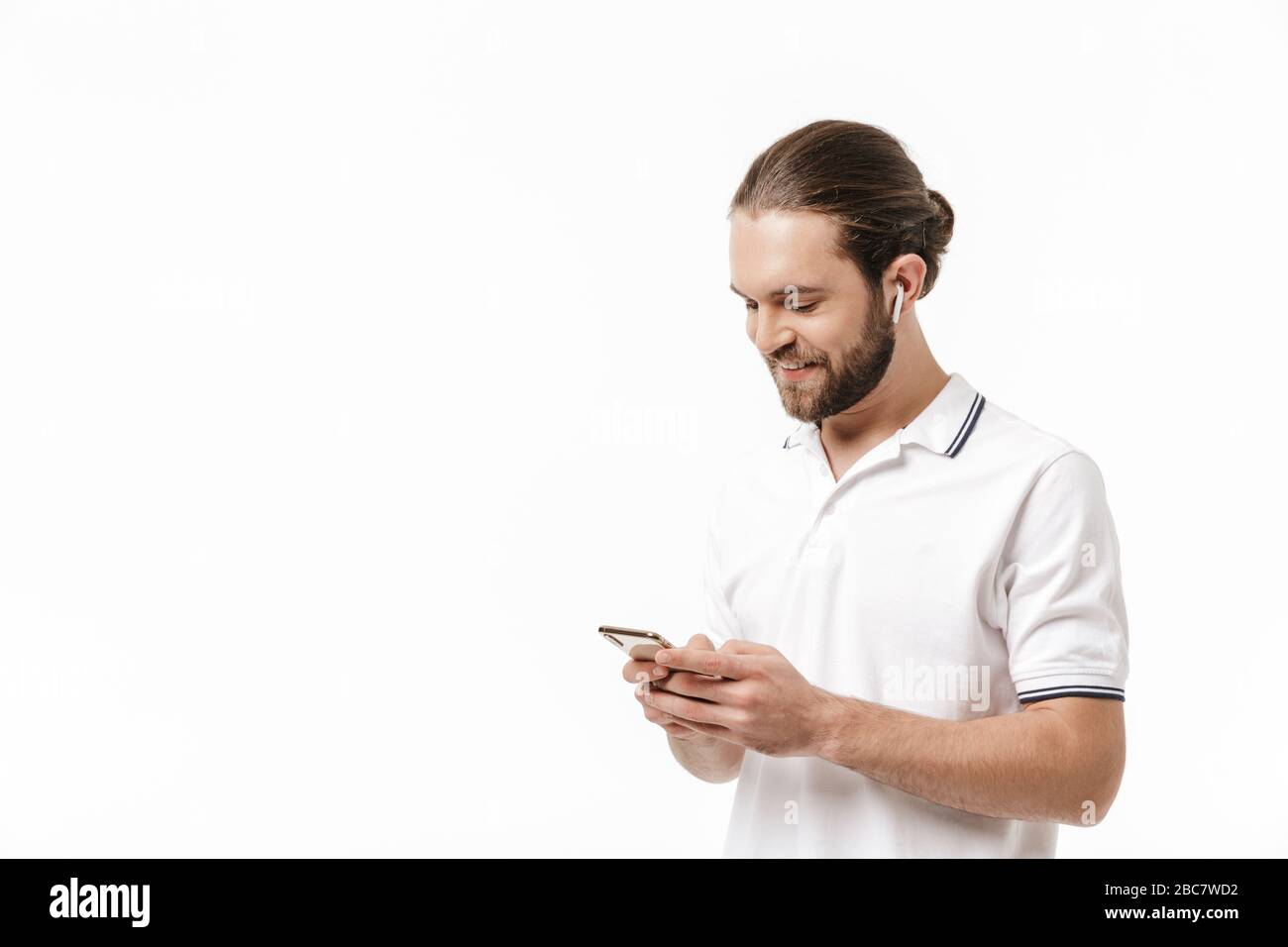 Foto di un giovane allegro positivo felice bel bell'uomo bearded che posa isolato su sfondo bianco muro utilizzando il telefono cellulare ascoltare musica con auricolare Foto Stock