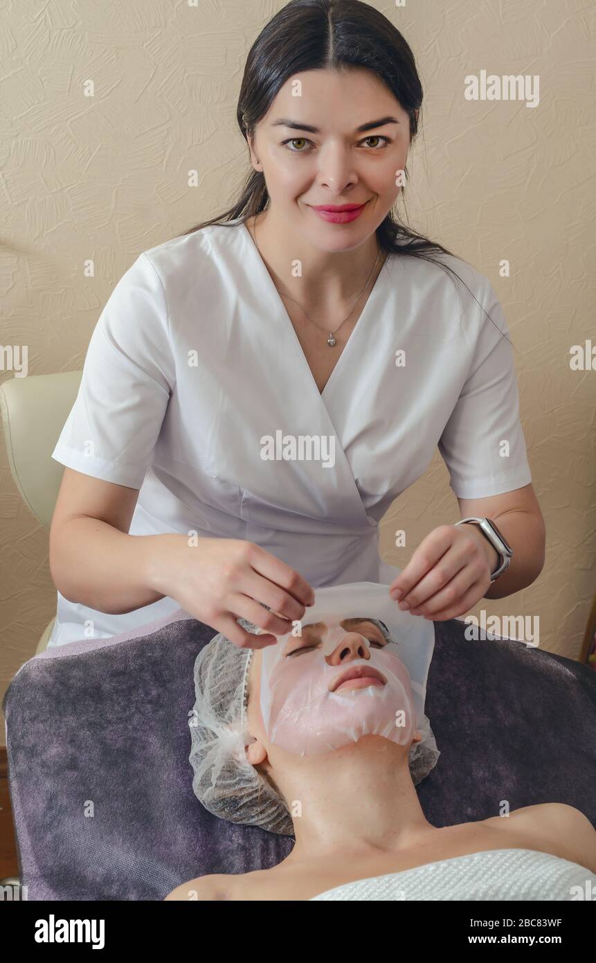 Beautician applicare la maschera di collagene alla faccia della donna. Cosmetologa lavora con una donna in un salone termale Foto Stock