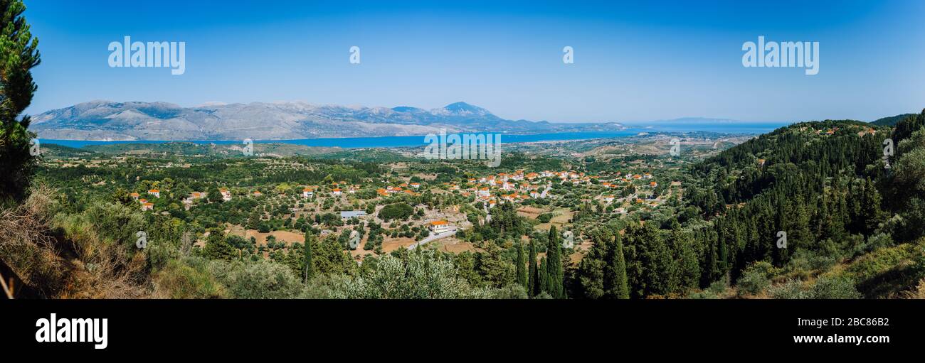 Vista panoramica ultra ampia dell'idilliaca cittadina a valle con tetti rossi sull'isola mediterranea. Oliveti, cipressi e baia blu in lontananza. Foto Stock