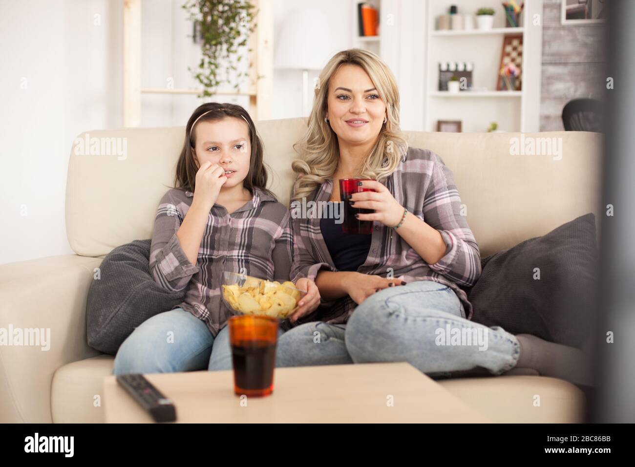 Madre e figlia che guardano un film seduto sul divano in una giornata pigra mangiando patatine e bevendo soda. Foto Stock