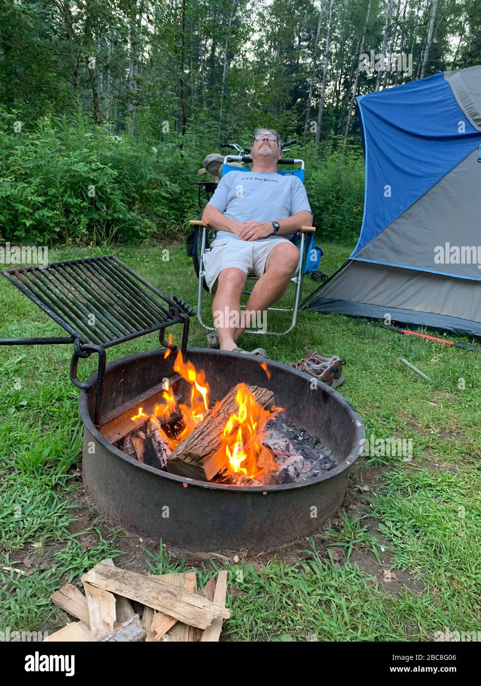 Un uomo di mezza età si sta rilassando davanti a un fuoco con la sua tenda sullo sfondo Foto Stock