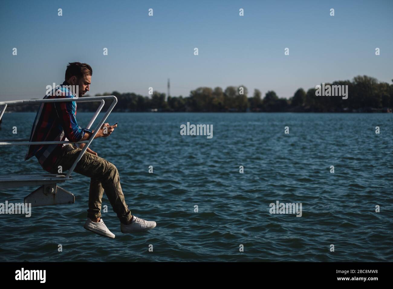 Serio bell'uomo che guarda il telefono cellulare mentre si siede sopra il fiume ona una bella giornata di sole Foto Stock