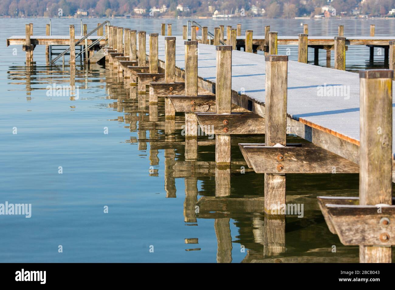 Vista lungo il lato di un molo in legno coperto di gelo a Starnberger See (Lago Starnberg). Con molte pile di legno e corrispondenti riflessi d'acqua. Foto Stock