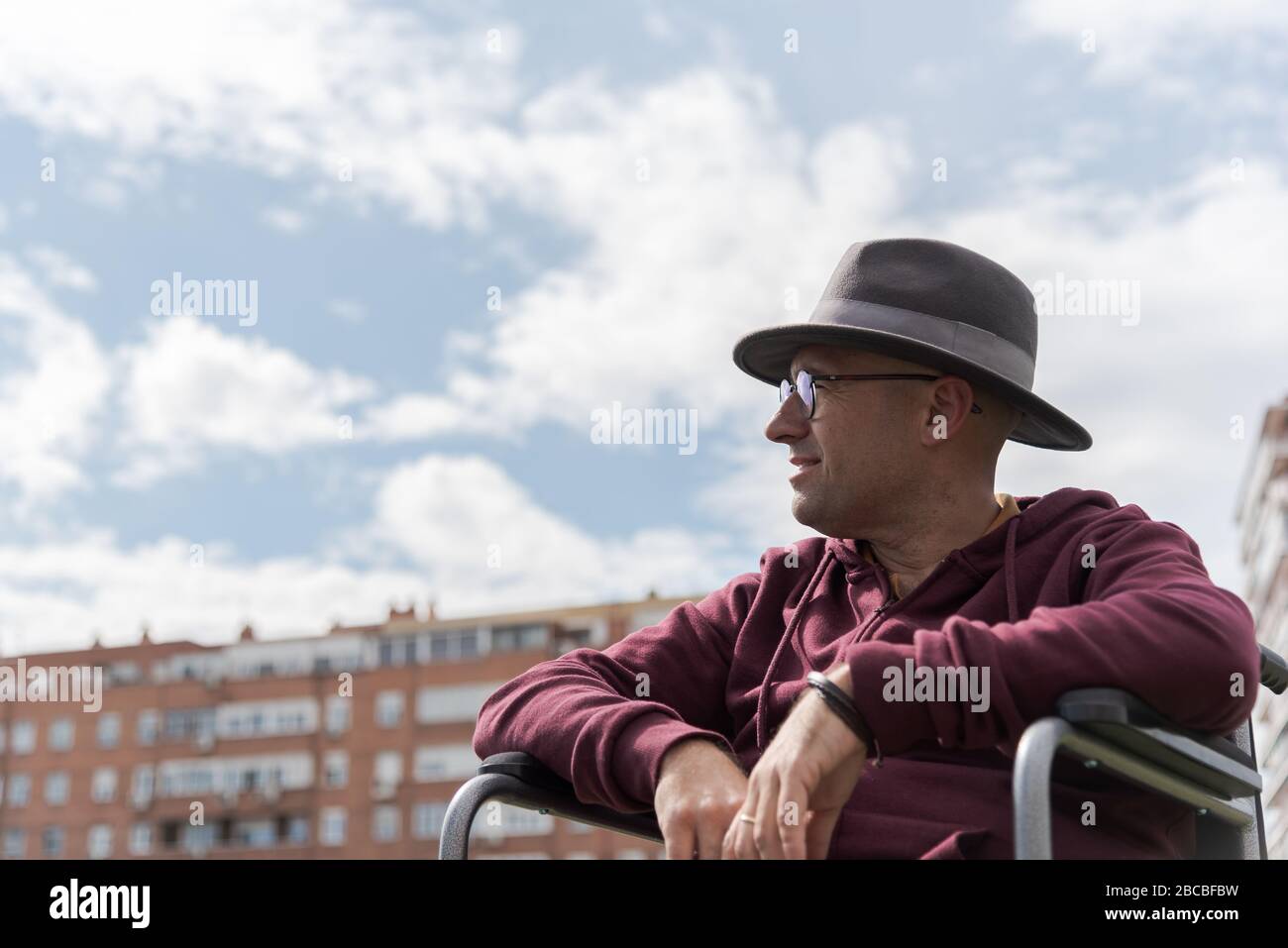 Uomo caucasico con occhiali e cappello depressivo e triste in una sedia a rotelle solitaria in un parco con il cielo e alcuni edifici sullo sfondo con lo spazio Foto Stock