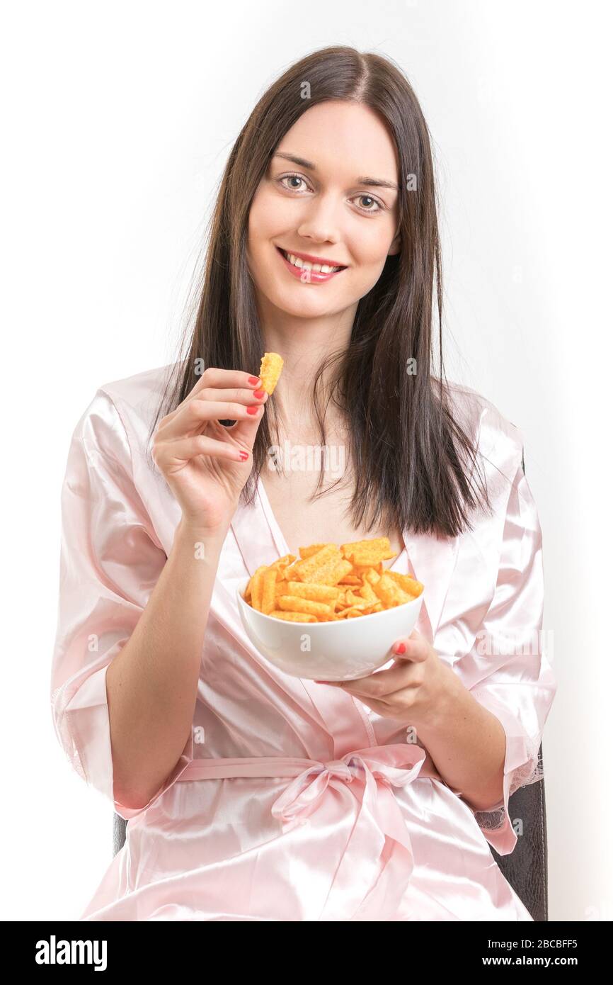 Bella giovane donna brunetta che indossa una abito da notte rosa, mangiare patatine sorridente. Foto di scorta senza royalty. Foto Stock