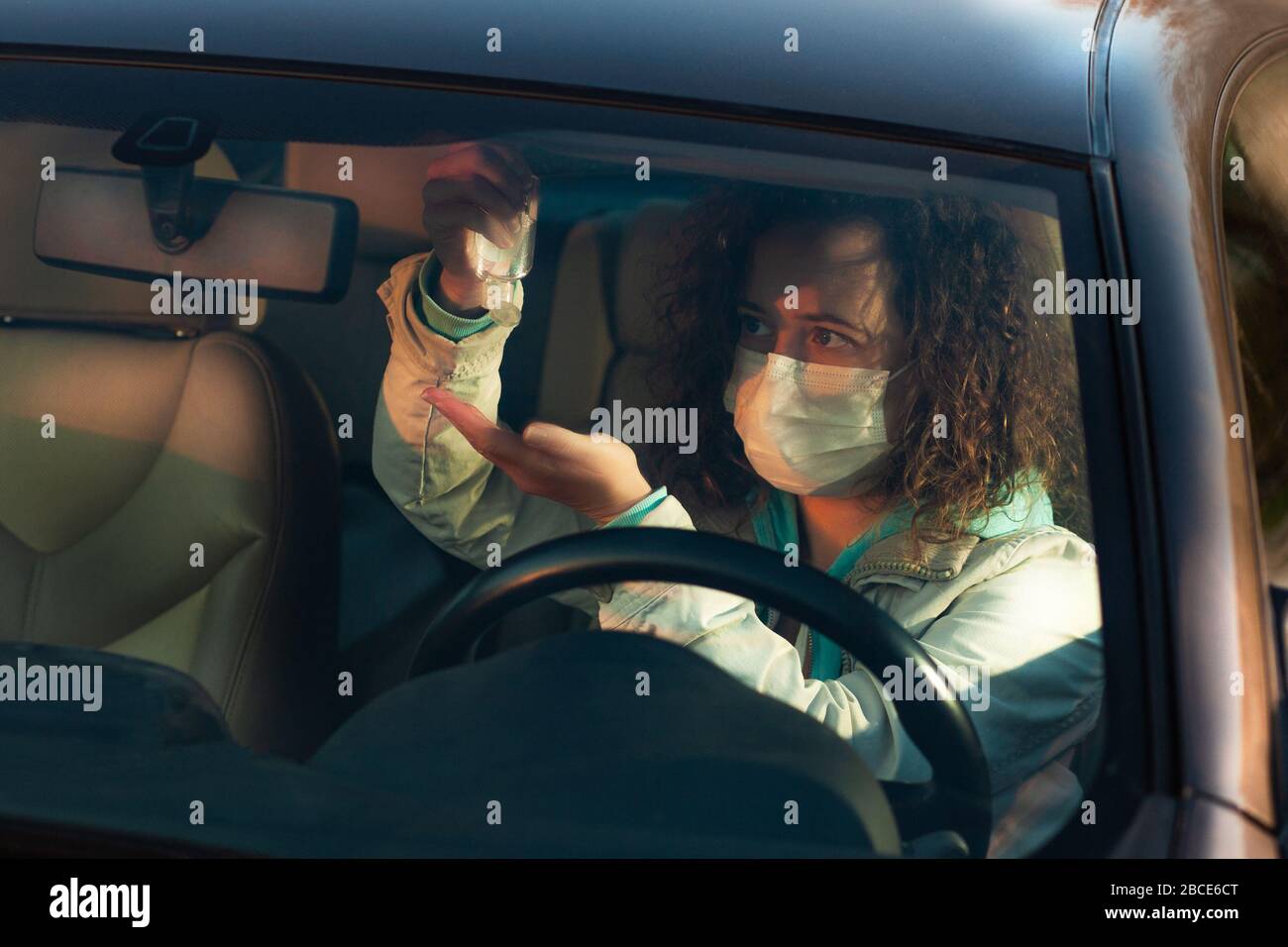 Donna curly che usa sanitizer nella sua automobile e che indossa la maschera medica nel traffico durante l'epidemia di covid-19 Foto Stock