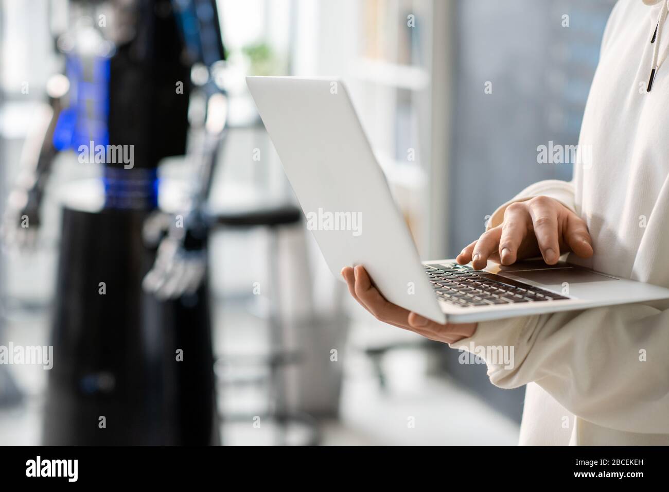 Mano di teenager con laptop che sviluppa nuovo software mentre lavora sopra progetto tecnologico sullo sfondo del robot nel college Foto Stock