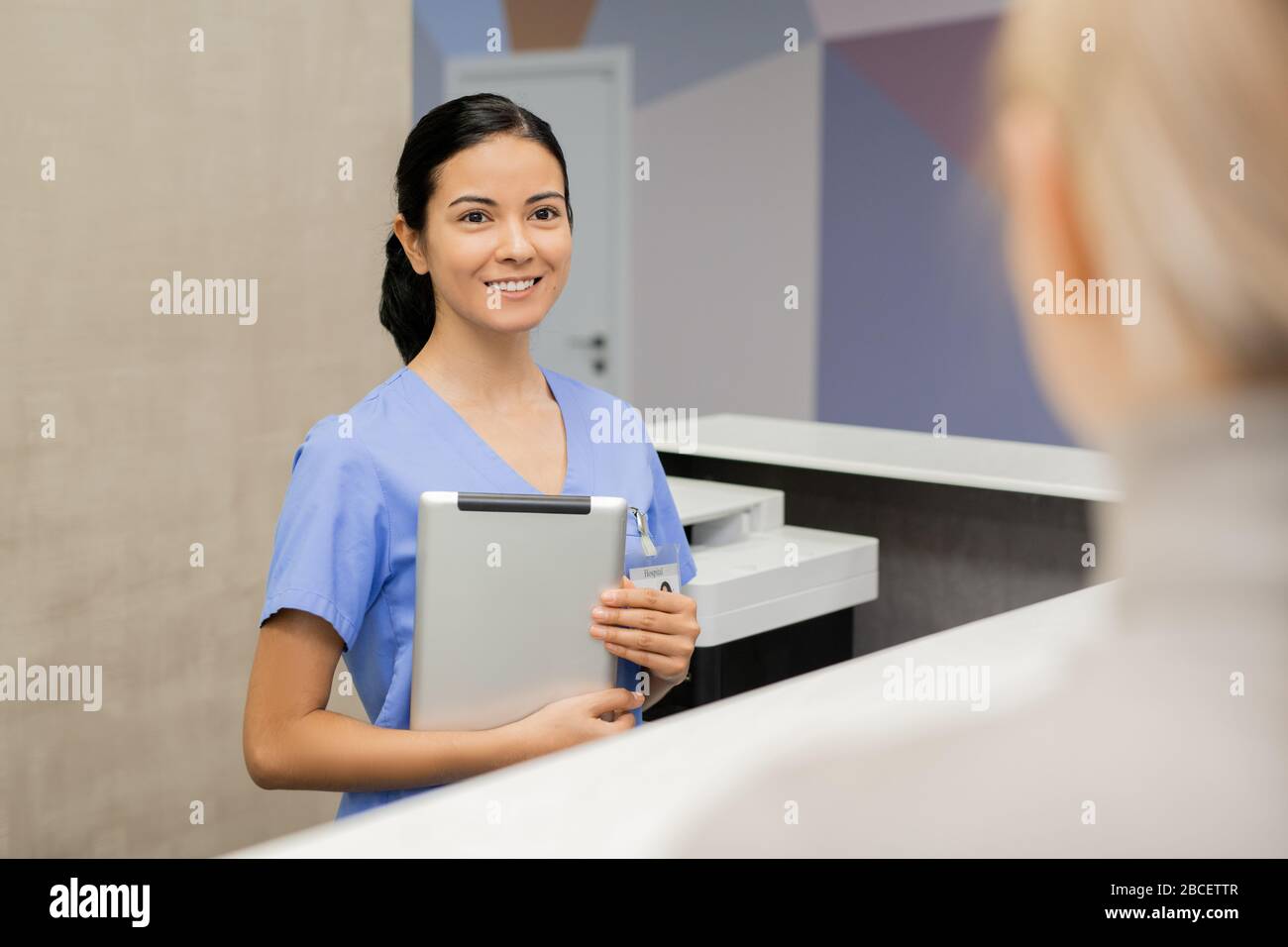 Allegro giovane amichevole assistente o receptionist in uniforme blu guardando il paziente di cliniche dentali mentre la consulta da banco Foto Stock