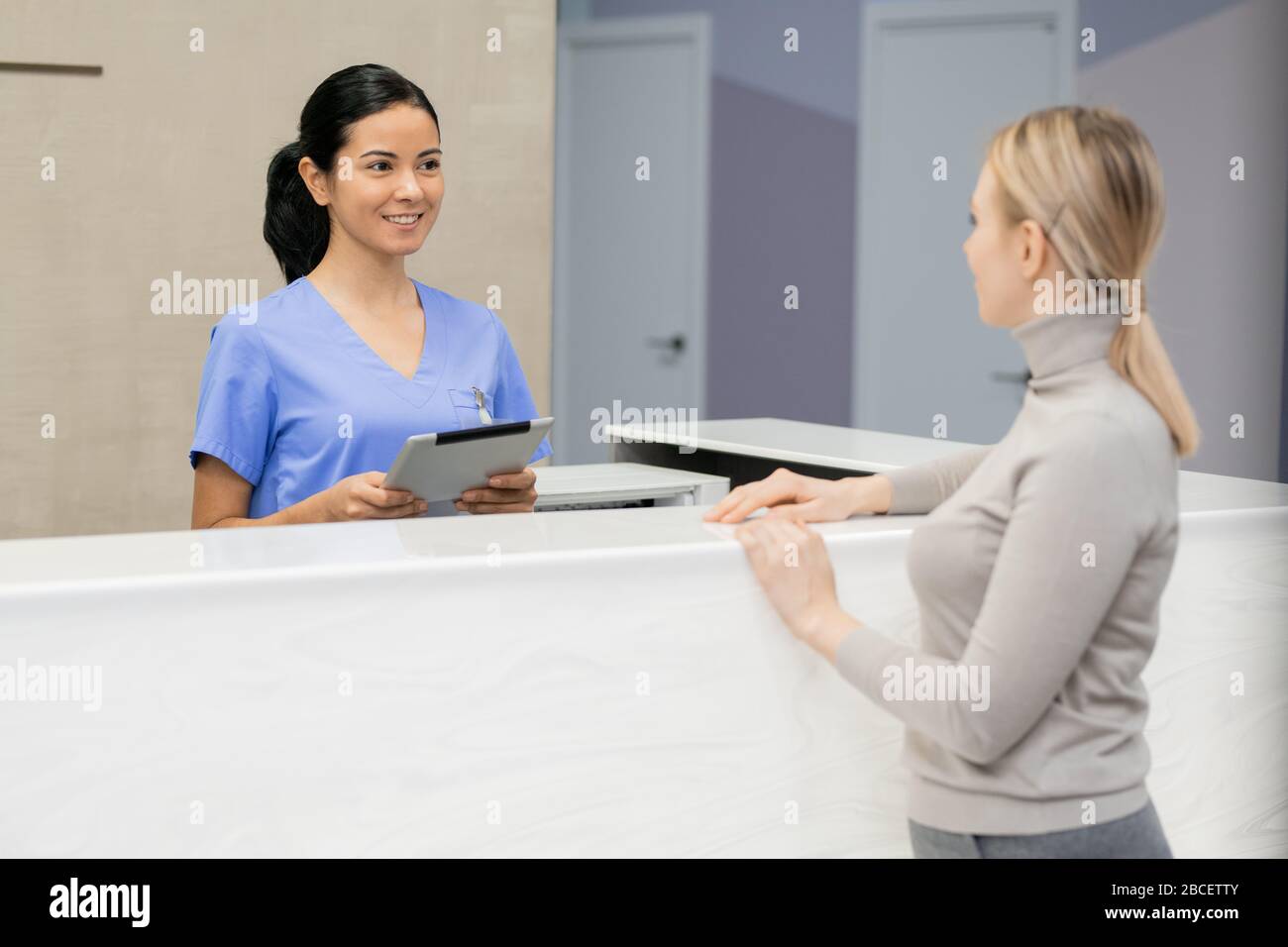 Consulente piuttosto giovane con la tavoletta in piedi dal banco di ricevimento di fronte alla paziente femminile e registrandola per l'appuntamento medico Foto Stock