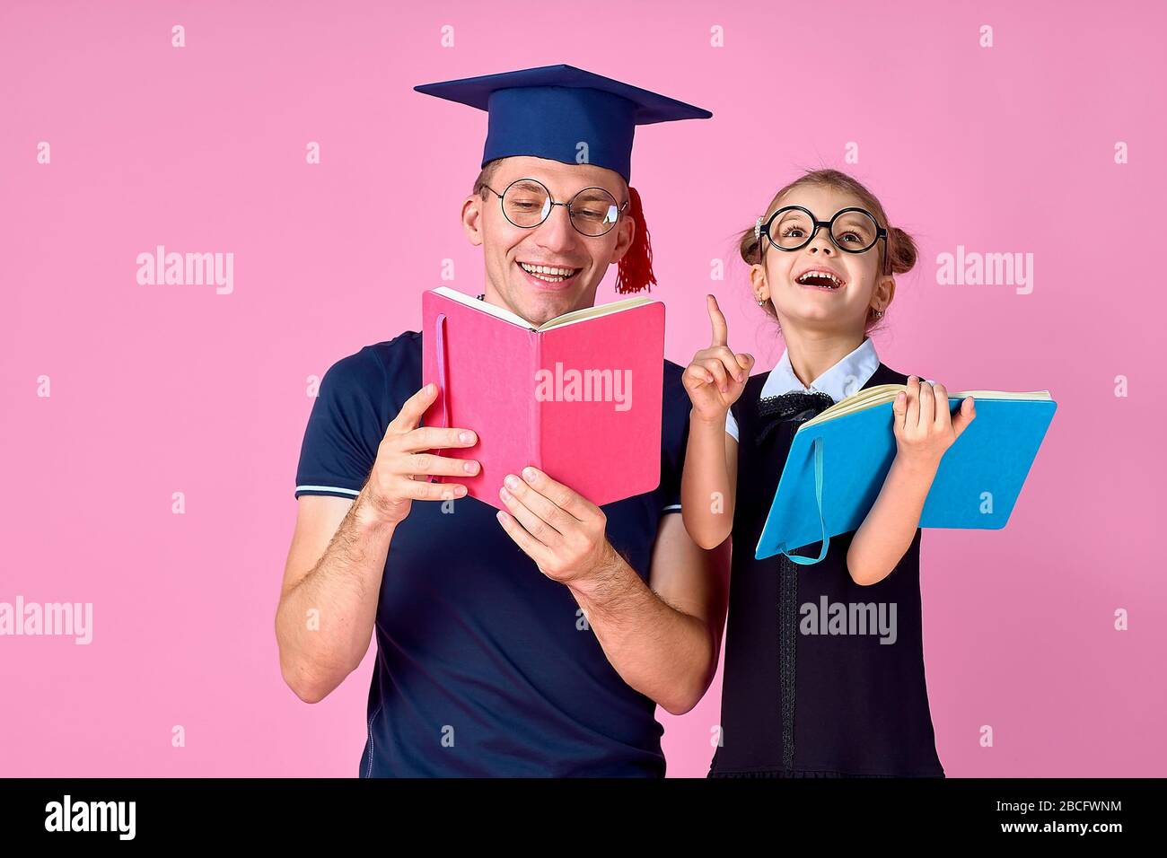 Uomo in cappello accademico che tiene il libro, studiare insieme con ragazza carina pre-teen in uniforme scolastica. Padre, figlia isolato su sfondo rosa in studio. Amore famiglia giorno concetto di infanzia genitorialità. Foto Stock