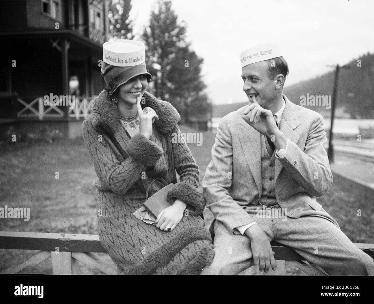 Un uomo e una donna che indossano cappelli divertenti si siedono a grinning  l'uno con le dita appuntate alla bocca, circa 1935. Il cappello della donna  recita 
