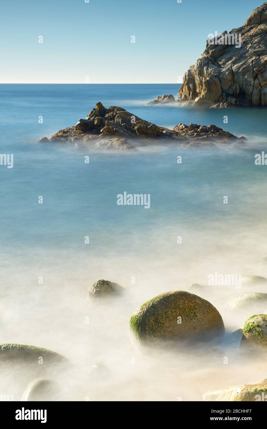 Spiaggia nascosta circondata da rocce modellate dall'acqua e il passaggio del tempo con piccole isolette colpite da un mare setoso, Blanes, Catalogna, Spagna. Foto Stock