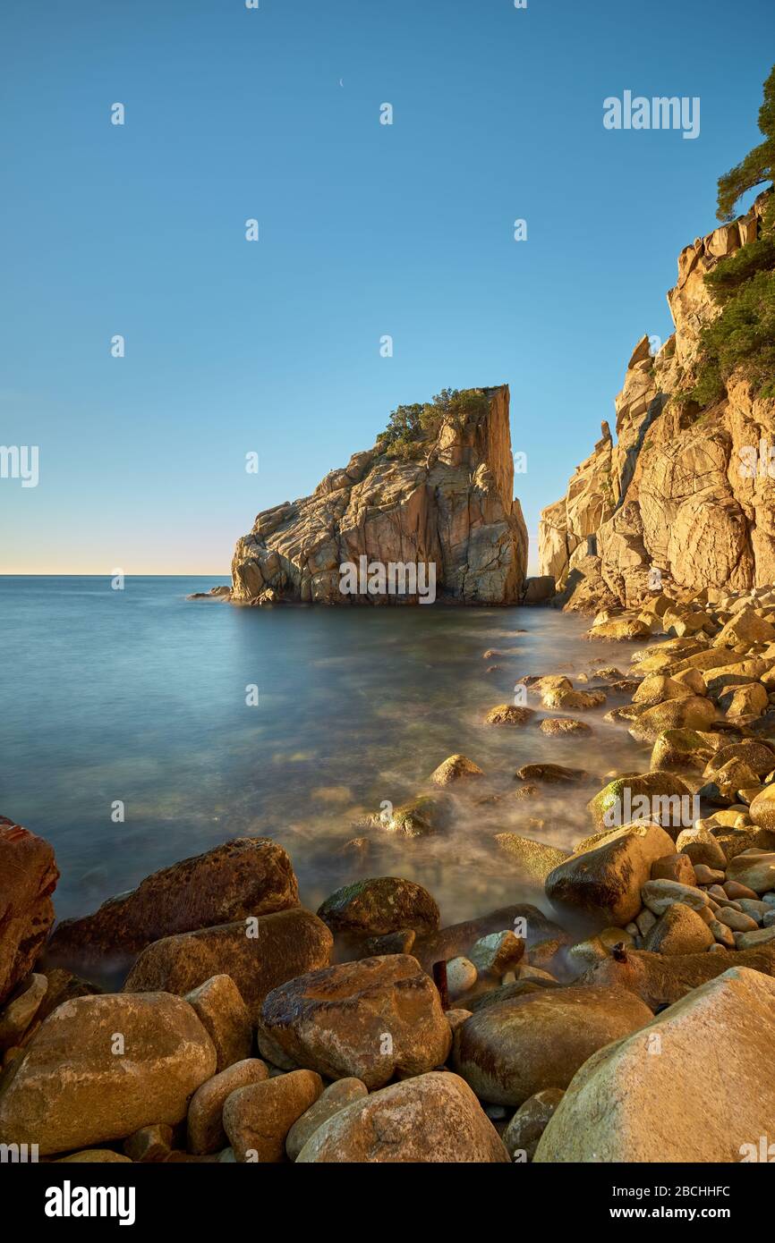 Rocce arrotondate dall'acqua sulla spiaggia e grande formazione rocciosa che aggettano nel mare in una giornata di sole, Blanes, Catalogna, Spagna. Foto Stock