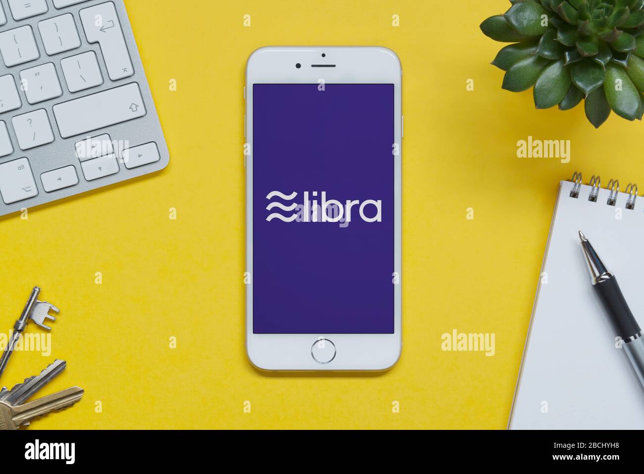 Un iPhone che mostra il logo di Facebook Libra si appoggia su una tabella di sfondo gialla con una tastiera, tasti, blocco note e impianto (solo per uso editoriale). Foto Stock