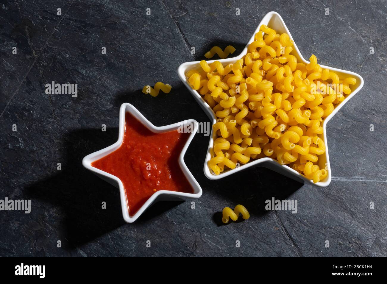Pasta secca e salsa di pomodoro in ciotole a forma di stella su un tavolo con sfondo di ardesia scura vista dall'alto, con spazio per le copie. Foto Stock
