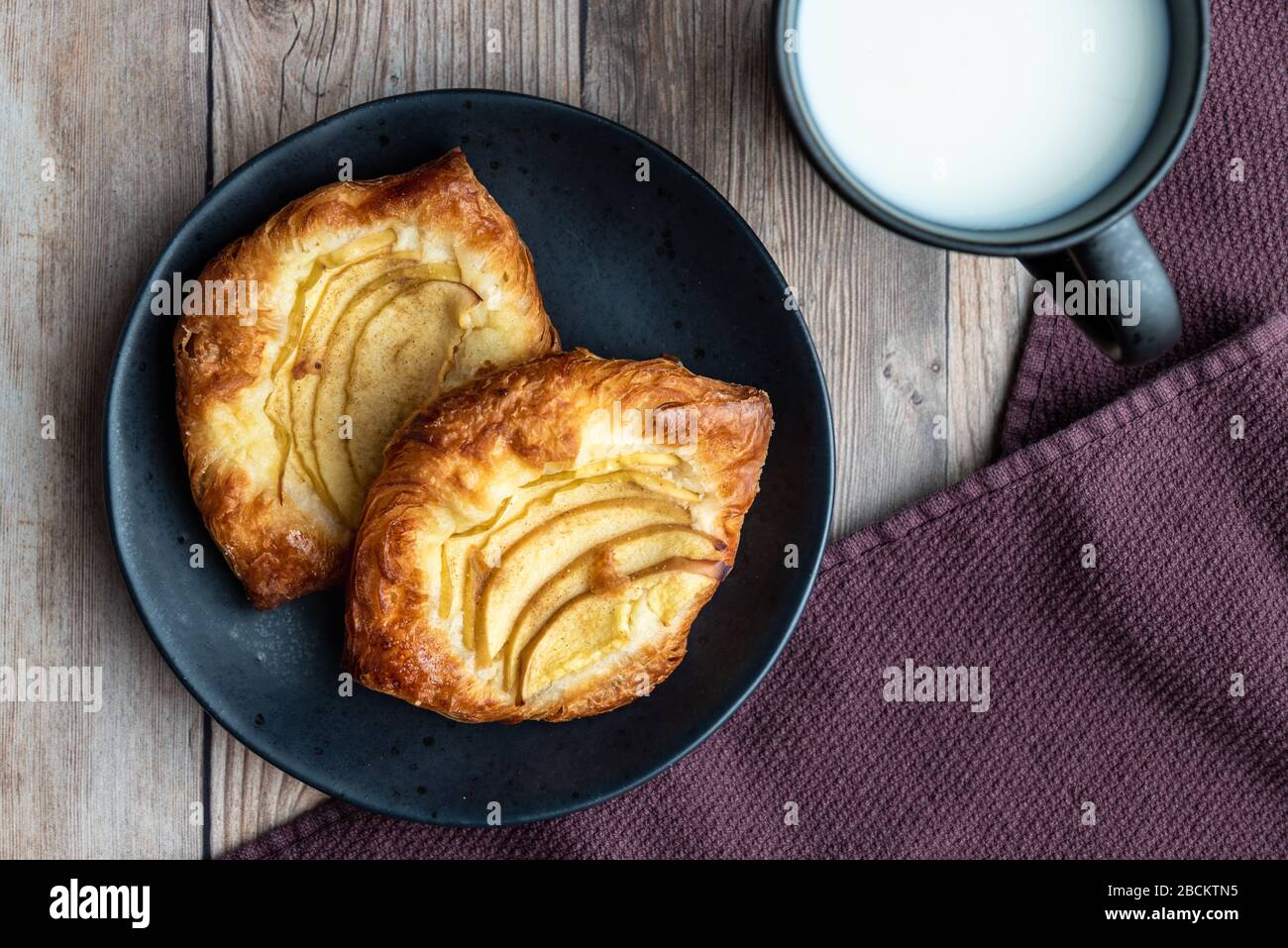 Crostate di mele singole fresche su un piatto nero, tazza di latte, tovagliolo di stoffa viola, sfondo di legno Foto Stock