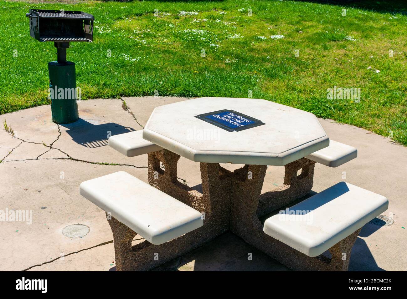 Tavolo da picnic in cemento nel parco pubblico con superficie non disinfettata cartello di avvertenza temporaneo attaccato al piano. Griglia in stile parco. Foto Stock