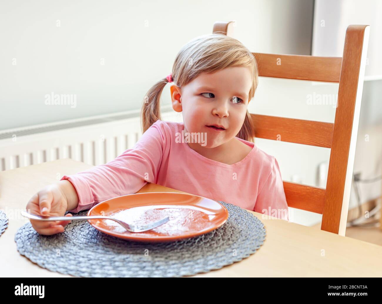 Bambina prescolare, finendo il suo pasto, seduto al tavolo su una sedia vicino ad un piatto vuoto che tiene una forchetta. Appena finito mangiare da sè Foto Stock
