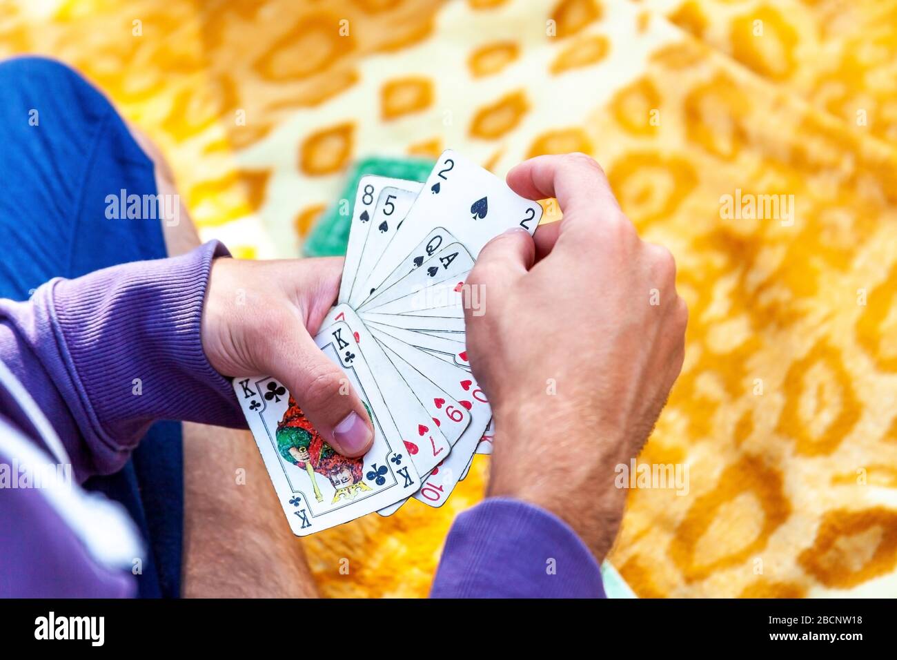 Giovane uomo in abbigliamento casual che gioca carte seduti su un blaket fuori, mani primo piano, la raccolta di una carta due di picche, tenendo un mazzo completo sparso fuori Foto Stock