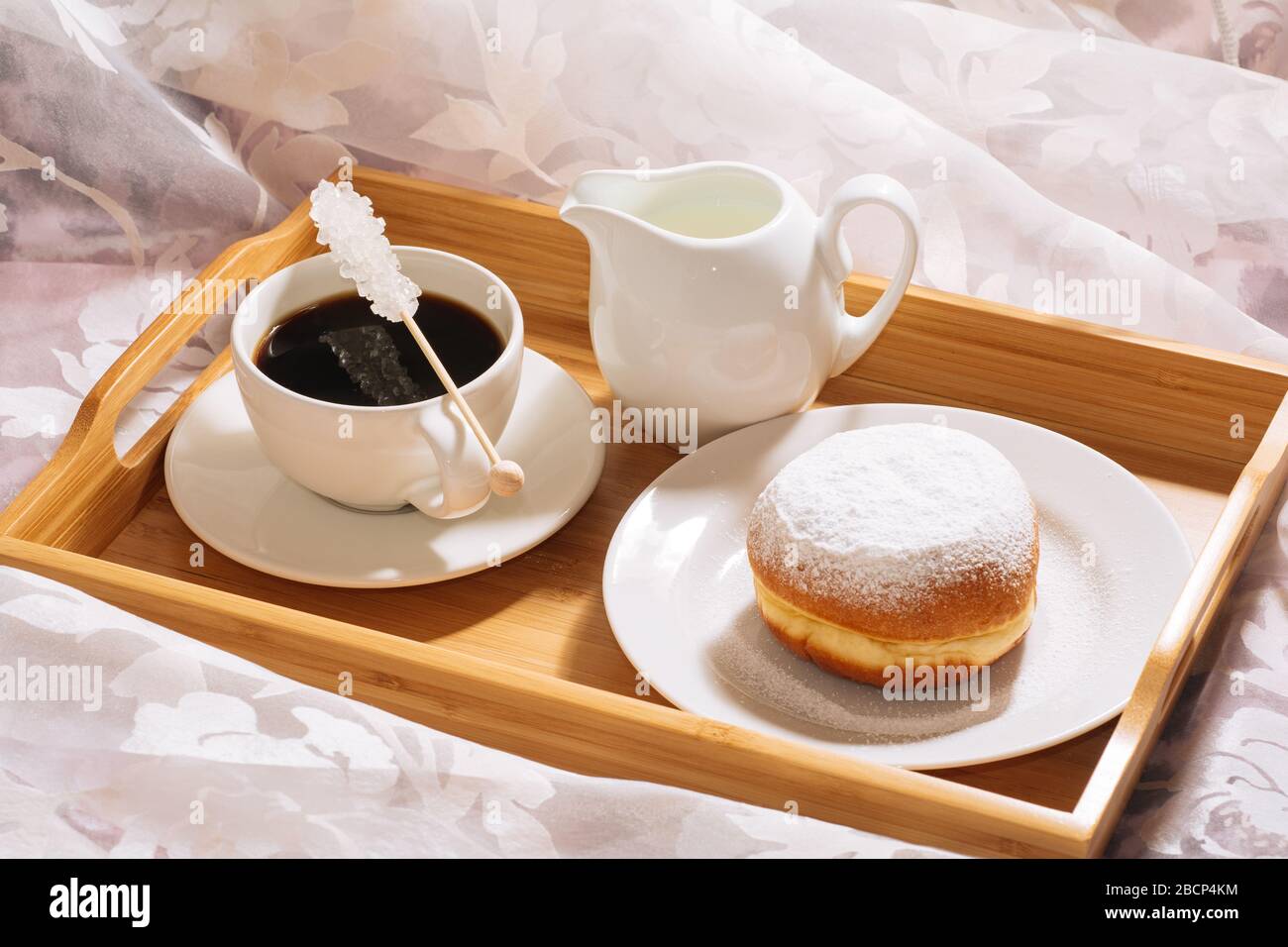 Brunch a letto con caffè appena tostato e caldo, caraffa di latte, ciambella in zucchero in polvere su un vassoio di legno in piatti bianchi. Orizzontale Foto Stock