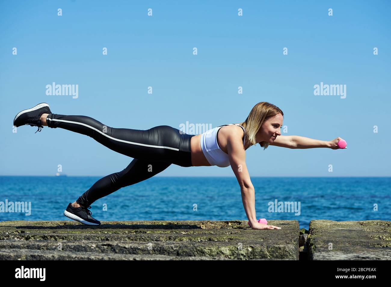 Foto orizzontale di una bella donna che fa esercizi sportivi sulla spiaggia dell'oceano o del mare. La donna sta tenendo manubri nelle sue mani. Foto Stock