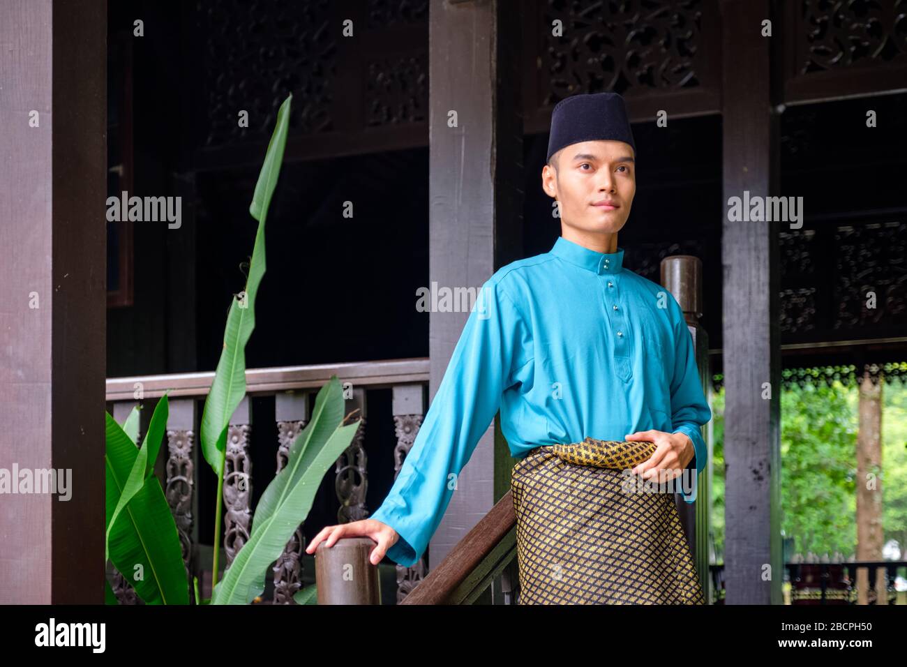 Un ritratto di giovane malese nella sua tradizionale stoffa teal, songket e songkok songkok a casa di legno tradizionale. Foto Stock