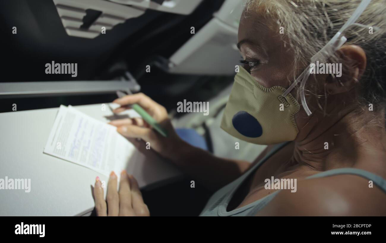 Donna viaggiare caucasico in aereo con indossare maschera medica protettiva. Ragazza turistica a bordo di un aereo con respiratore di protezione, compilare un modulo di registrazione. Foto Stock