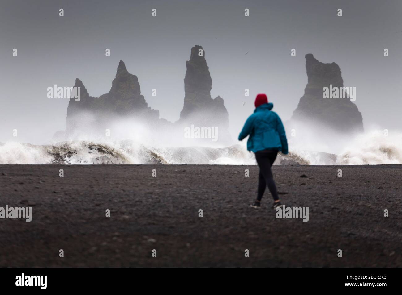 Ragazza che cammina sulla spiaggia di sabbia nera con i Reynisdrangar Seastacks sullo sfondo, Vik, Islanda Foto Stock