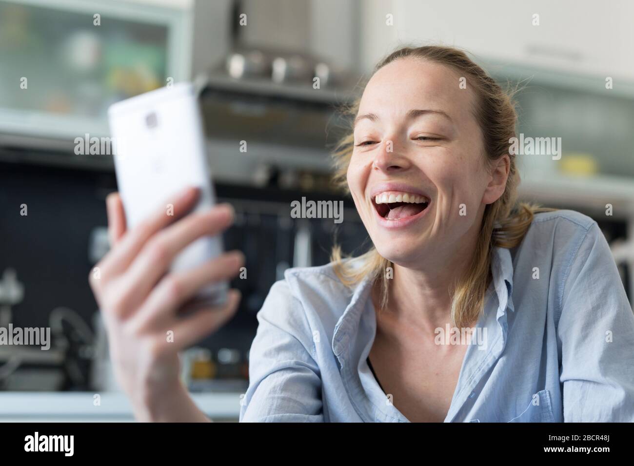 Giovane sorridente allegra donna felice al chiuso in cucina a casa utilizzando le app di social media sul telefono cellulare per chattare e stare in contatto con la sua amata Foto Stock