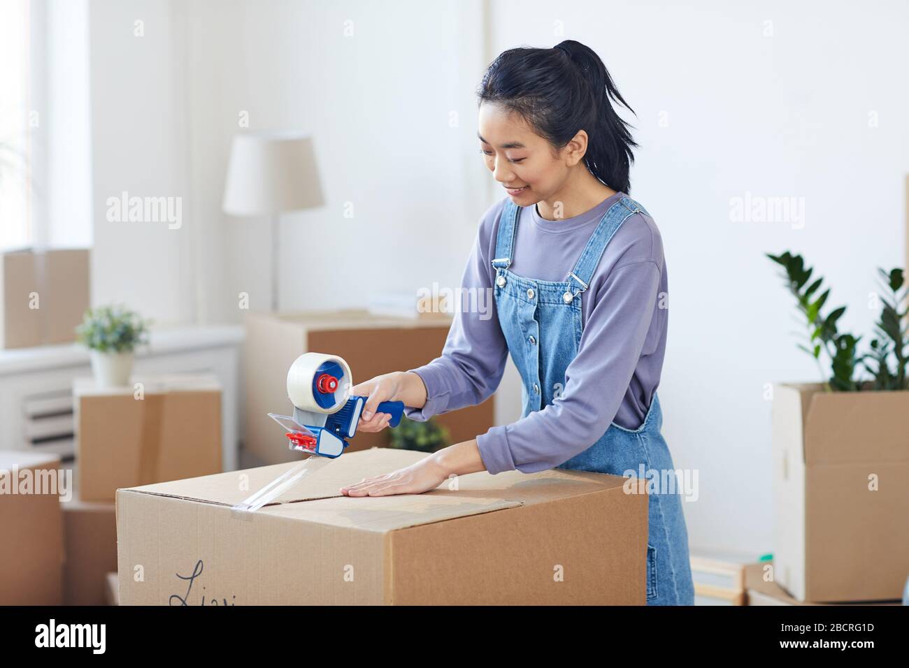 Ritratto di donna asiatica sorridente imballaggio scatole di cartone con dispenser di nastro mentre st si sposta verso casa nuova, copia spazio Foto Stock