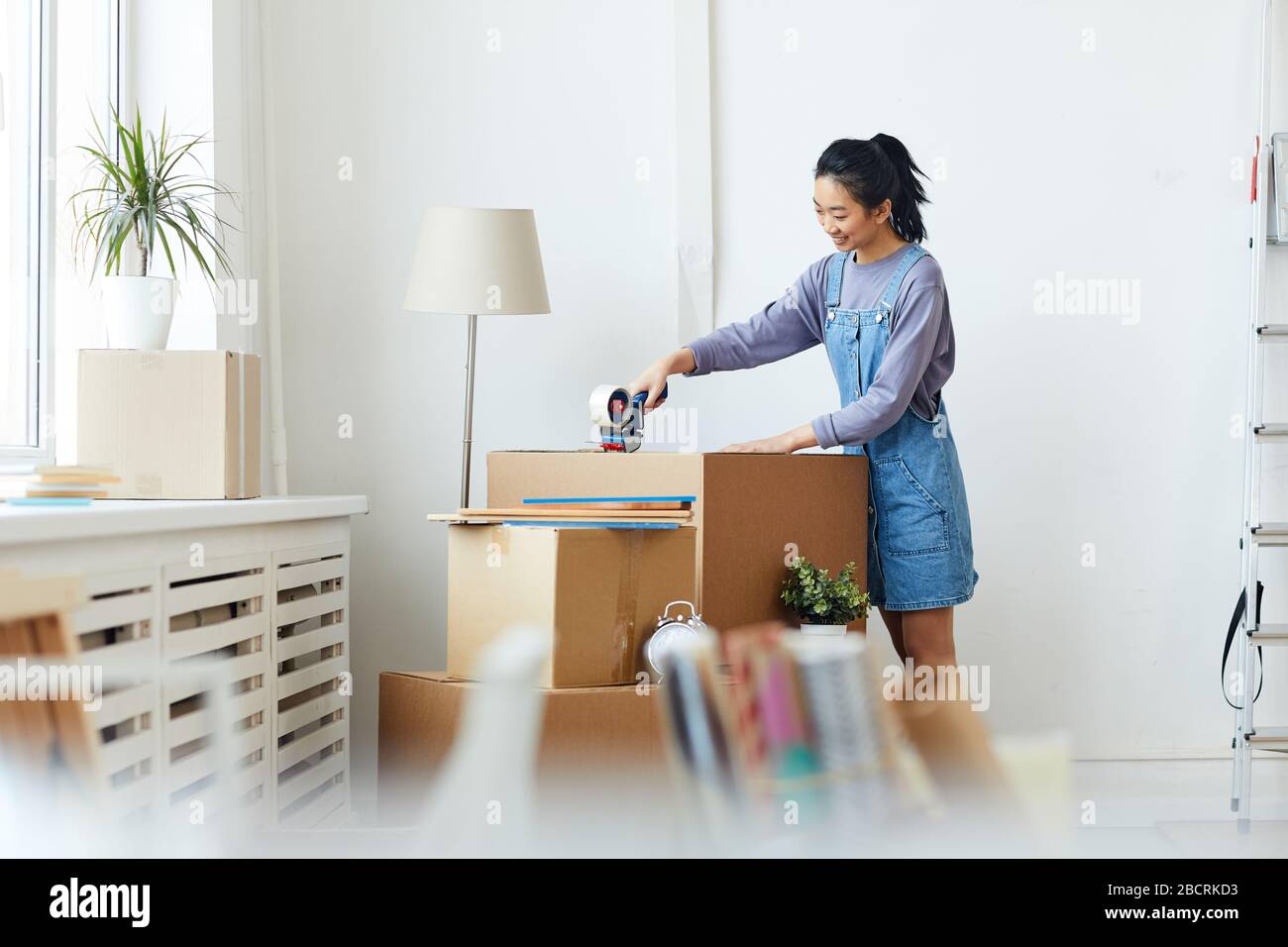 Vista laterale ritratto di giovane donna asiatica imballaggio scatole di cartone e sorridente felice mentre si sposta a casa nuova o appartamento, copia spazio Foto Stock