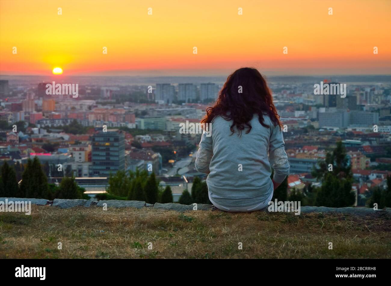 Da sola, giovane ragazza seduta sopra la città e che guarda al calmo tramonto estivo. Foto Stock