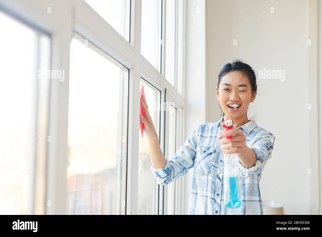 Girovita ritratto di giovane donna asiatica che punta la pistola a spruzzo alla fotocamera mentre si lava le finestre e godendo la pulizia della primavera, copia spazio Foto Stock