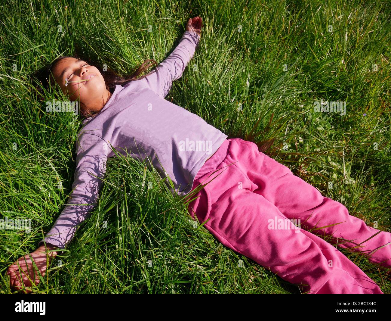 Bambina sorridente e rilassata, distesa sull'erba, con un fiore in bocca. Vista dall'alto Foto Stock