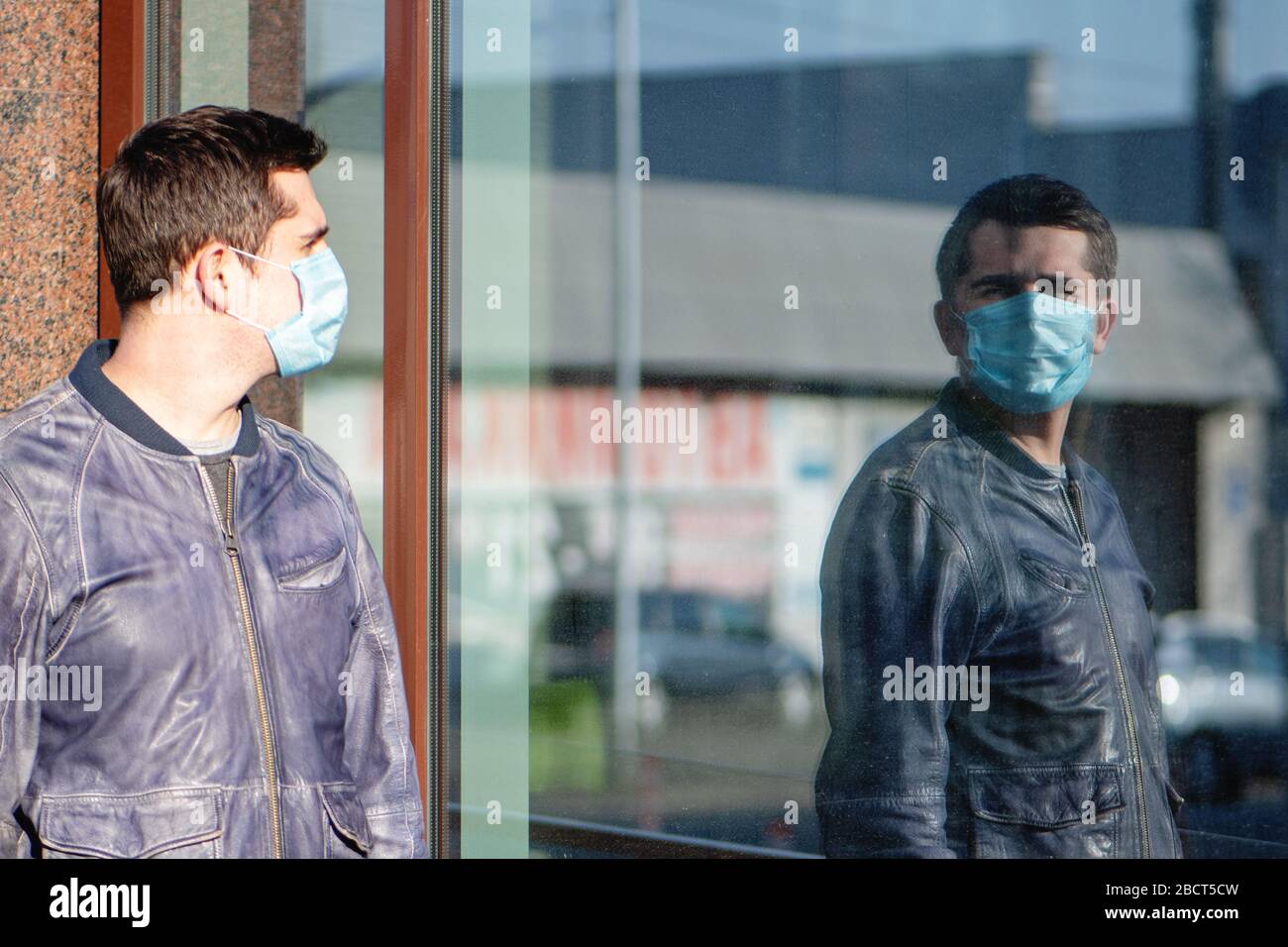 Giovane uomo in maschera medica protettiva guardando il suo riflesso nella finestra della strada. Foto Stock