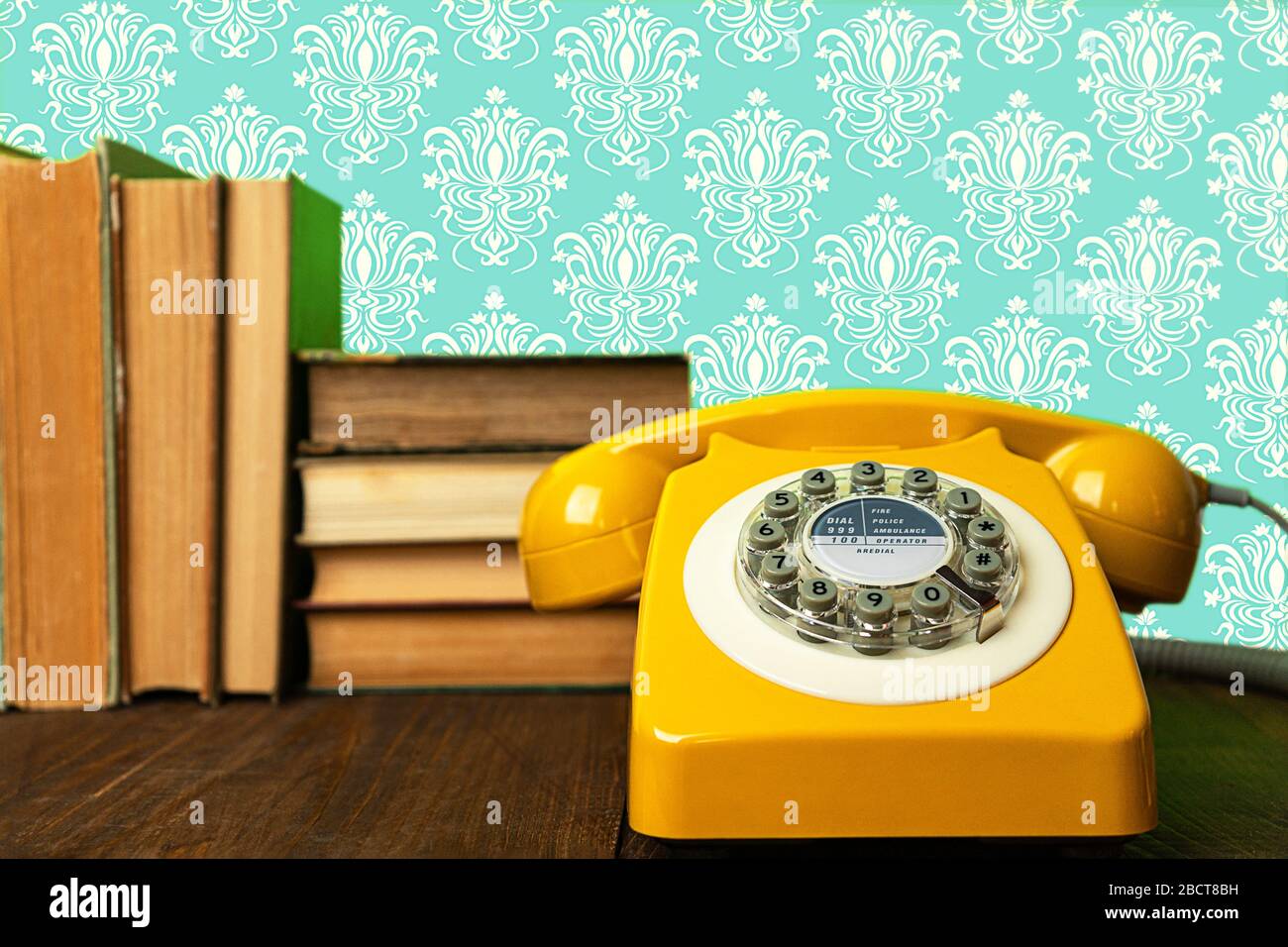 Telefono retrò giallo su tavolo di legno con vecchi libri e sfondo astratto con motivi geometrici. Foto Stock