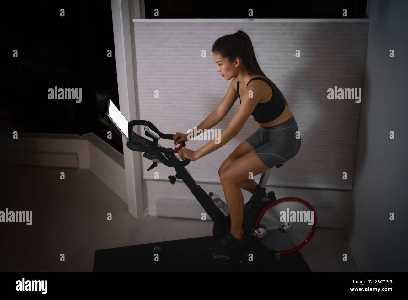 Home workout indoor cyclette Asian girl bike screen con lezioni online donna allenamento su attrezzature fitness smart indoor per l'esercizio in bicicletta. A tarda notte in camera da letto. Foto Stock