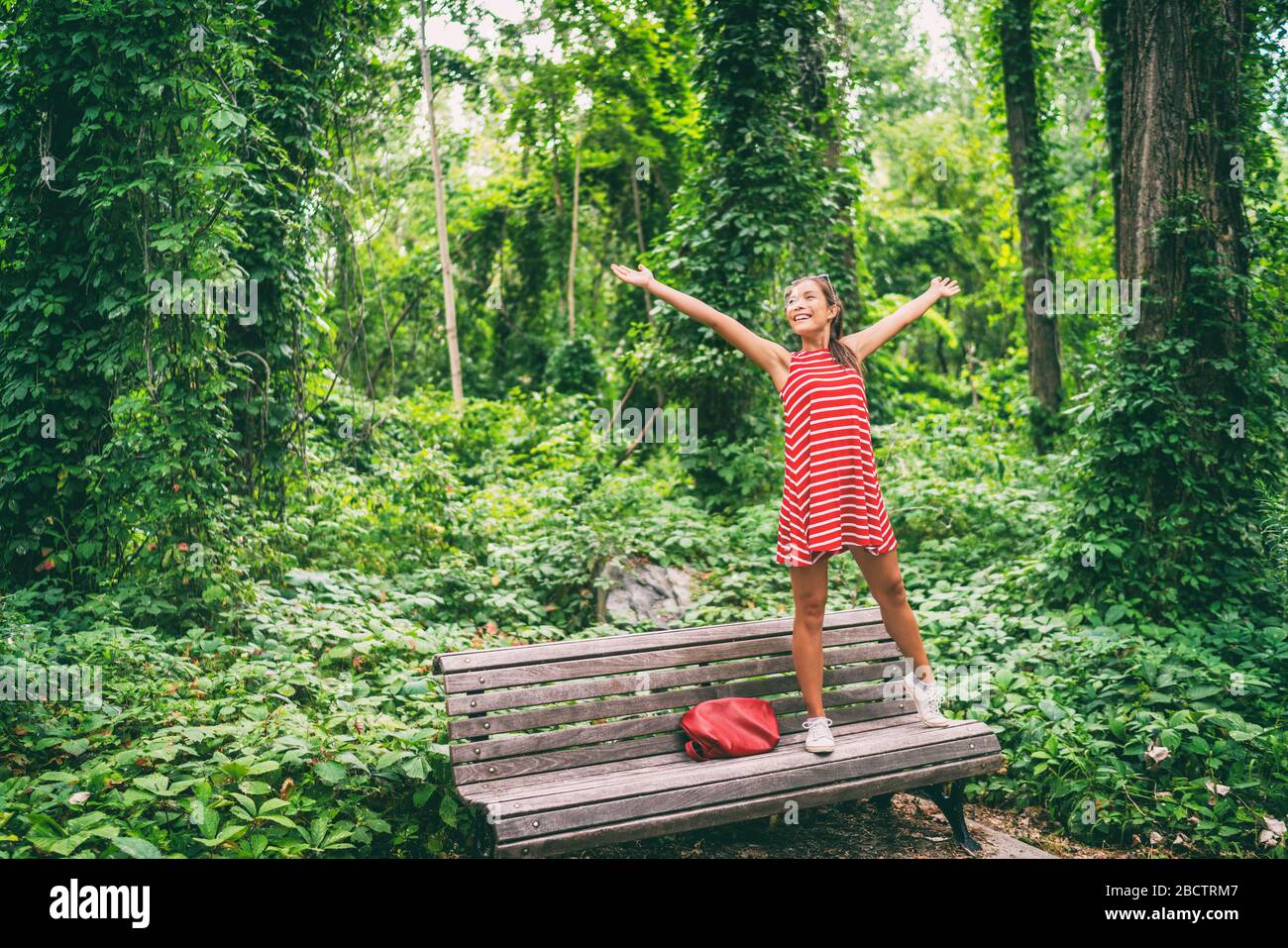 Felice donna asiatica godendo di libertà in estate natura alberi in piedi su panca parco con le braccia distese in gioia. Foto Stock