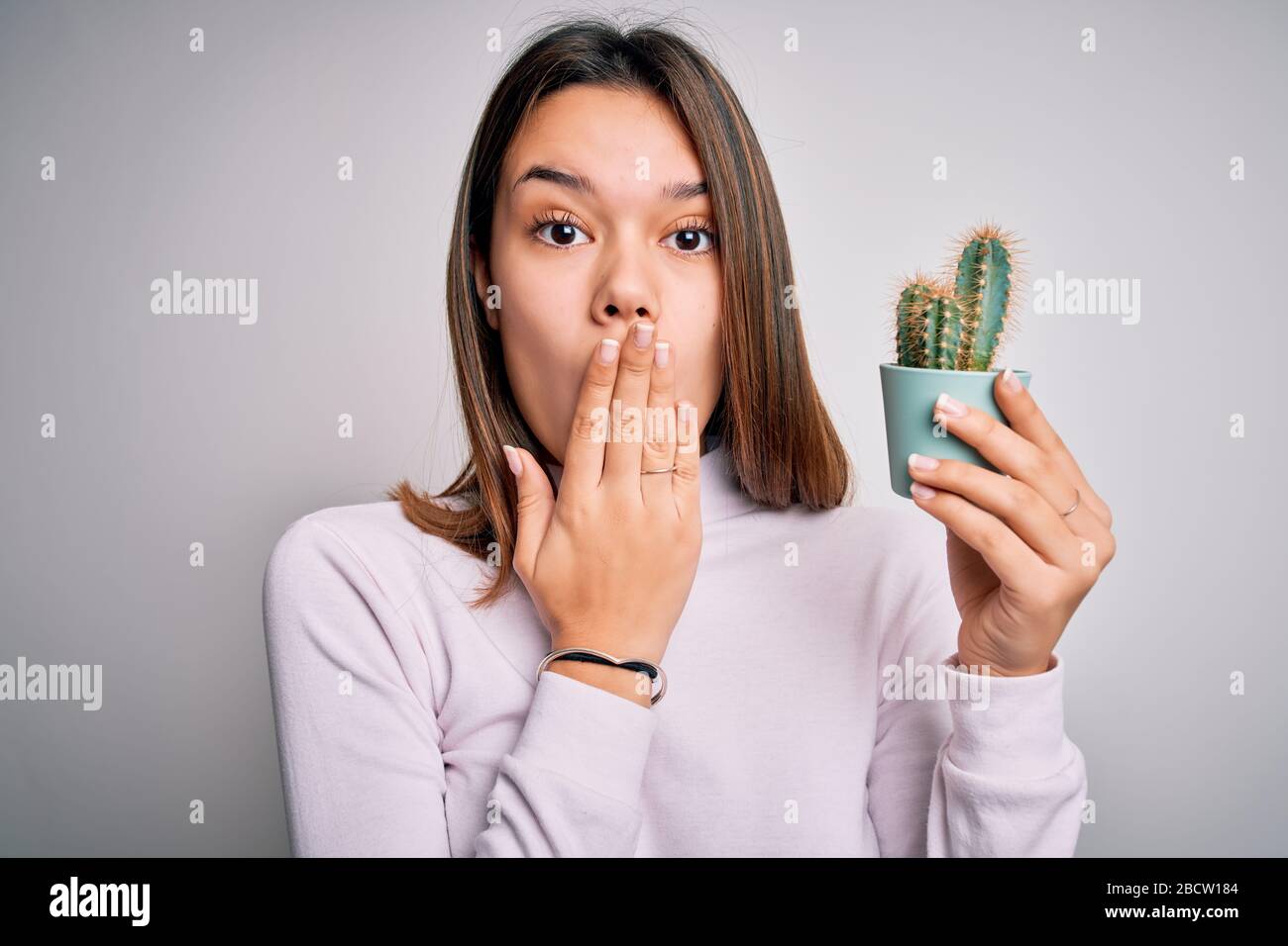 Giovane bella ragazza bruna che tiene piccolo vaso di pianta di cactus sopra la bocca di copertura bianca isolata di fondo con la mano scioccata con vergogna per errore, expr Foto Stock